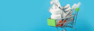 Are International Pharmacies Safe - Online Pharmacy Shopping - Miniature silver shopping cart filled with white medication bottles, blister packs, and syringes on a blue background.