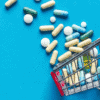 Cost Plus Drugs Alternative: Better Options for Cheap Medications - International Online Pharmacy - Miniature shopping cart filled with various capsules and medication pills spilled on a blue background.